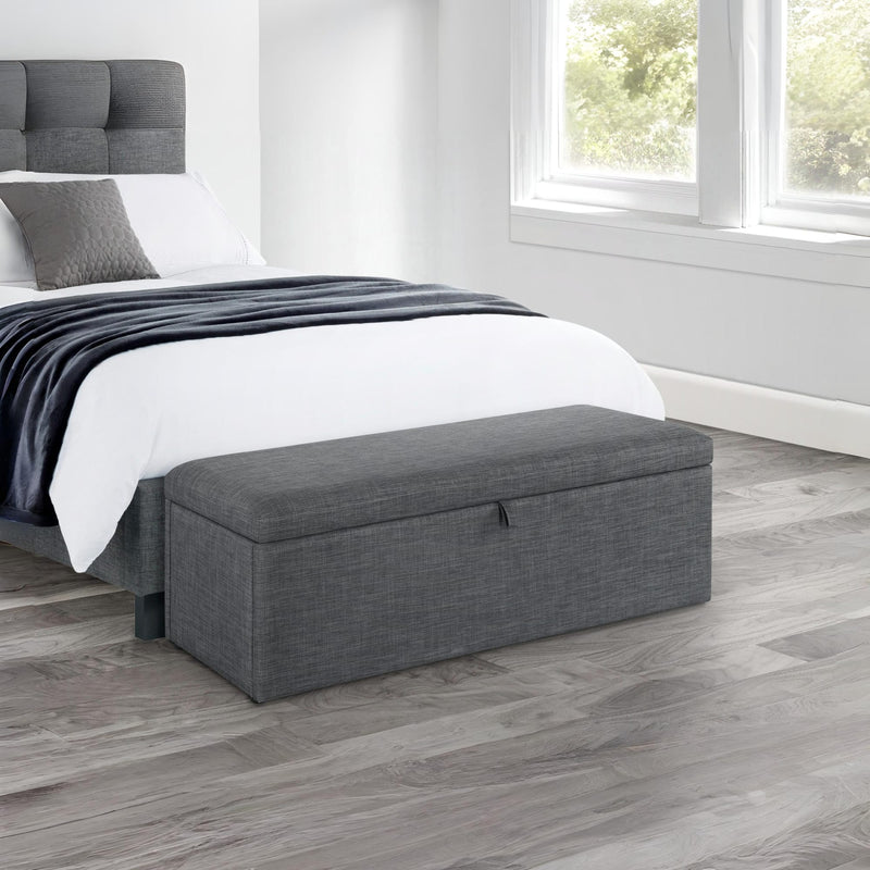 Gray storage bench in front of a bed in a bedroom with wooden flooring.