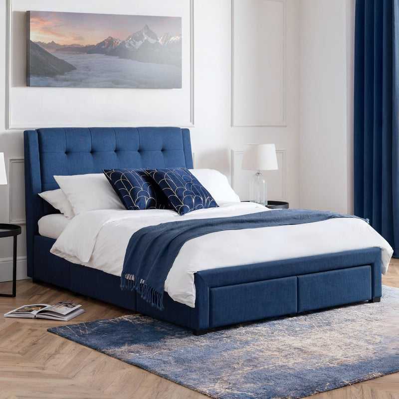 Blue upholstered bed with white bedding in a room with a mountain painting and lamp.