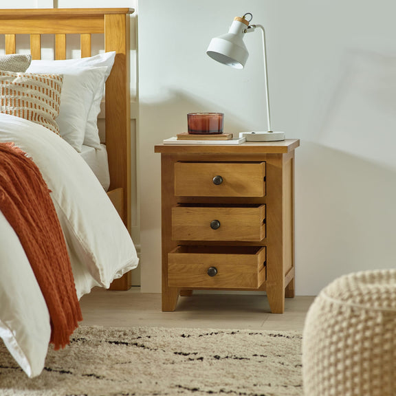 Wooden nightstand with three drawers next to a bed in a bedroom setting.