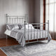 Silver metal bed with bedding in a room with wooden flooring and curtains.