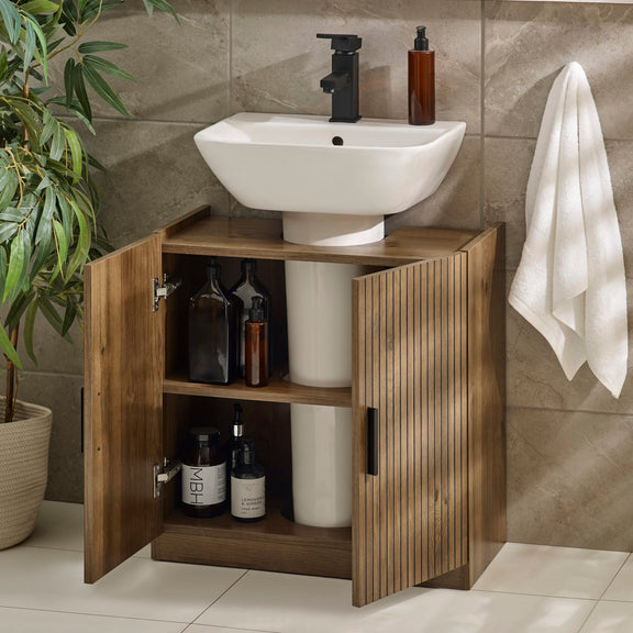 Bathroom setup with a wooden cabinet under a white sink, bottles inside, and a towel on a wall.