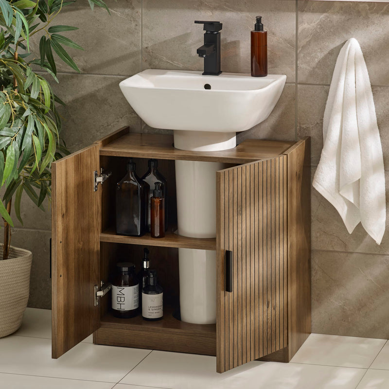 Bathroom setup with a wooden cabinet under a white sink, bottles inside, and a towel on a wall.