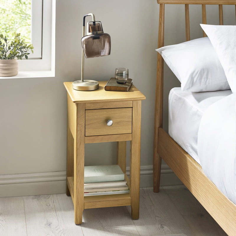 Wooden bedside table with lamp and books next to a bed in a bedroom.