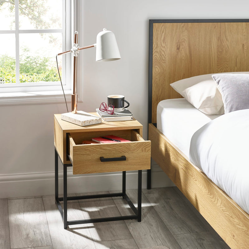 Wooden nightstand with books and a cup next to a bed in a bright bedroom.