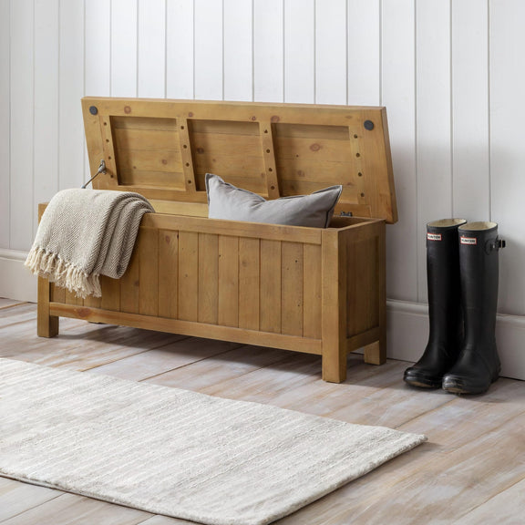 Wooden storage bench with a blanket and pillow, black boots nearby on a wooden floor.