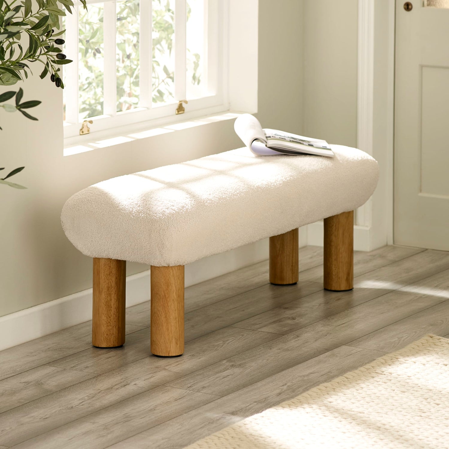 Beige ottoman with wooden legs in a room with a window and plant.