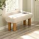 Beige ottoman with wooden legs in a room with a window and plant.