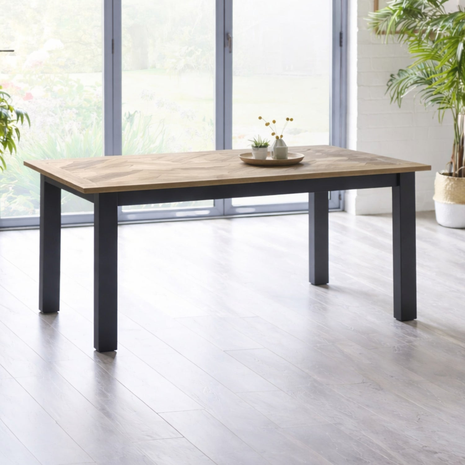 Wooden dining table with black legs in a bright room with large windows.