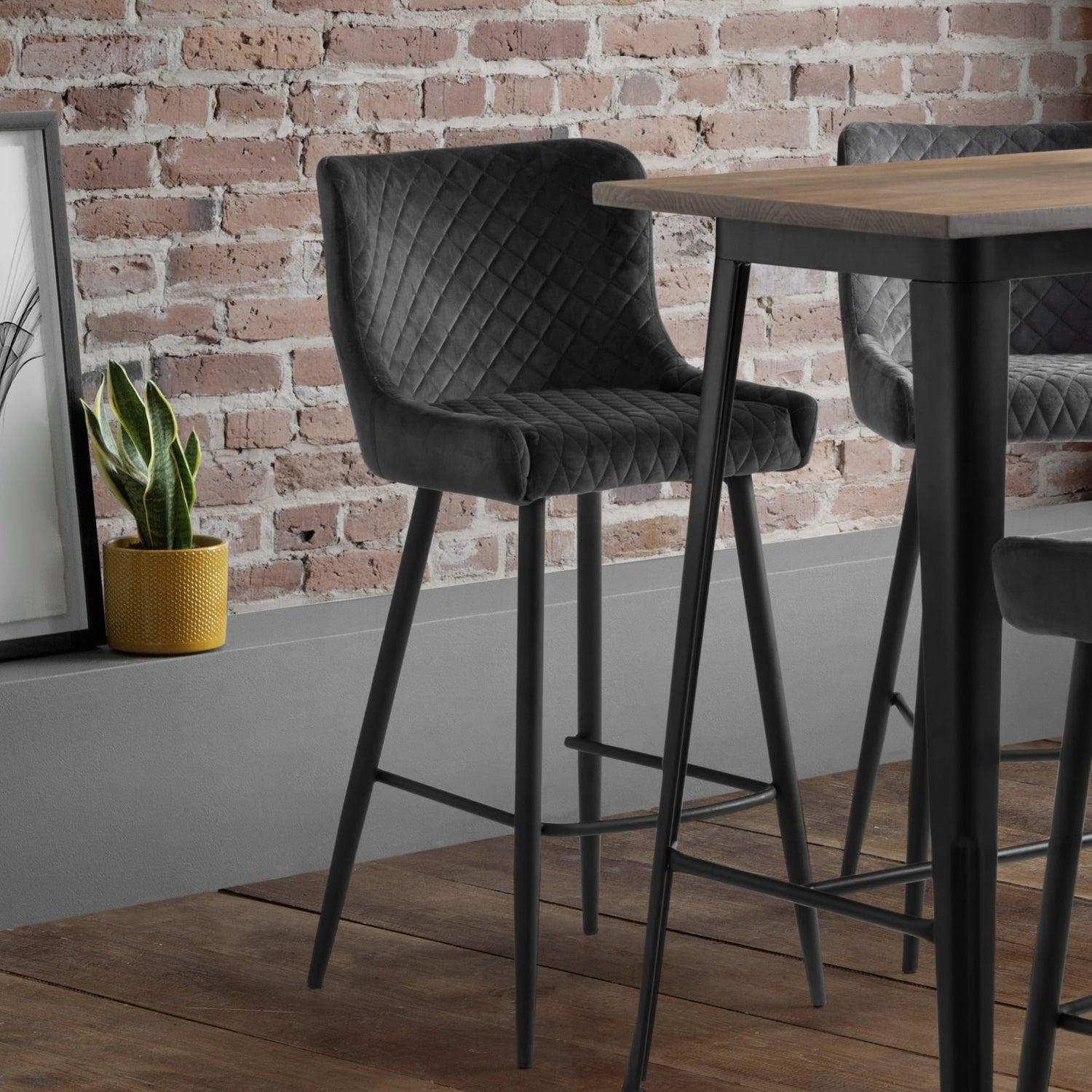 Gray quilted bar stools in front of a wooden table against a brick wall.