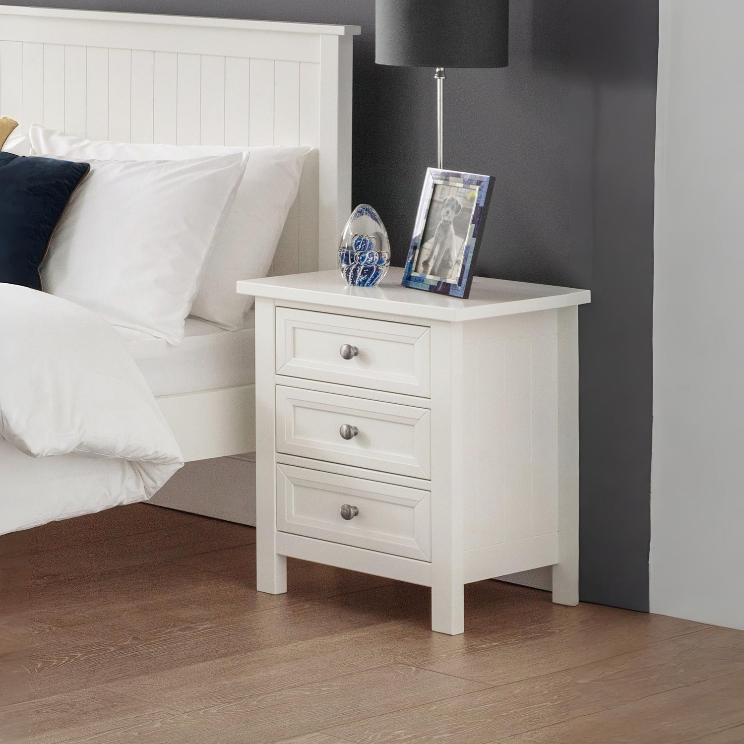 White nightstand with decorative items next to a bed with white bedding and a dark headboard.