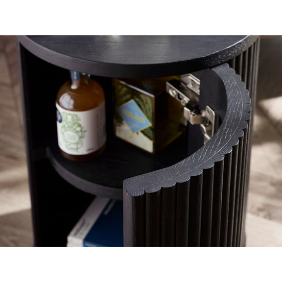 Black cylindrical side table with shelves holding a bottle and books, on a wooden floor.