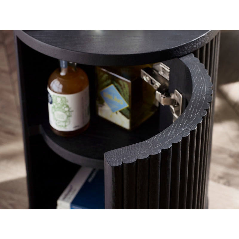 Black cylindrical side table with shelves holding a bottle and books, on a wooden floor.