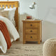 Wooden nightstand with three drawers next to a bed with white bedding and an orange throw blanket.