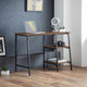 Wooden desk with a laptop, camera, and books in a room with gray walls and wooden floor.