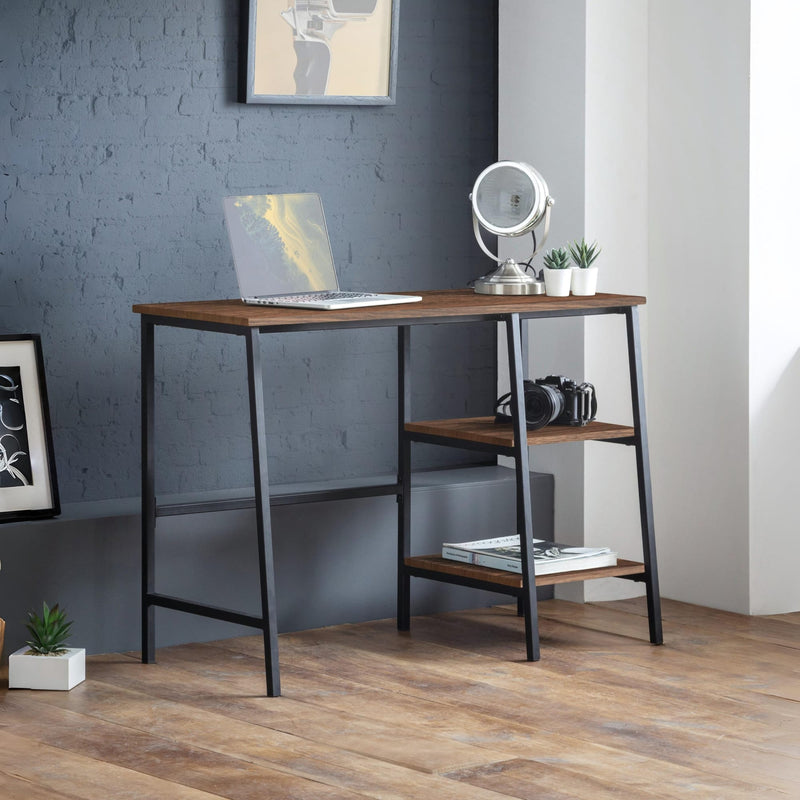 Wooden desk with a laptop, camera, and books in a room with gray walls and wooden floor.