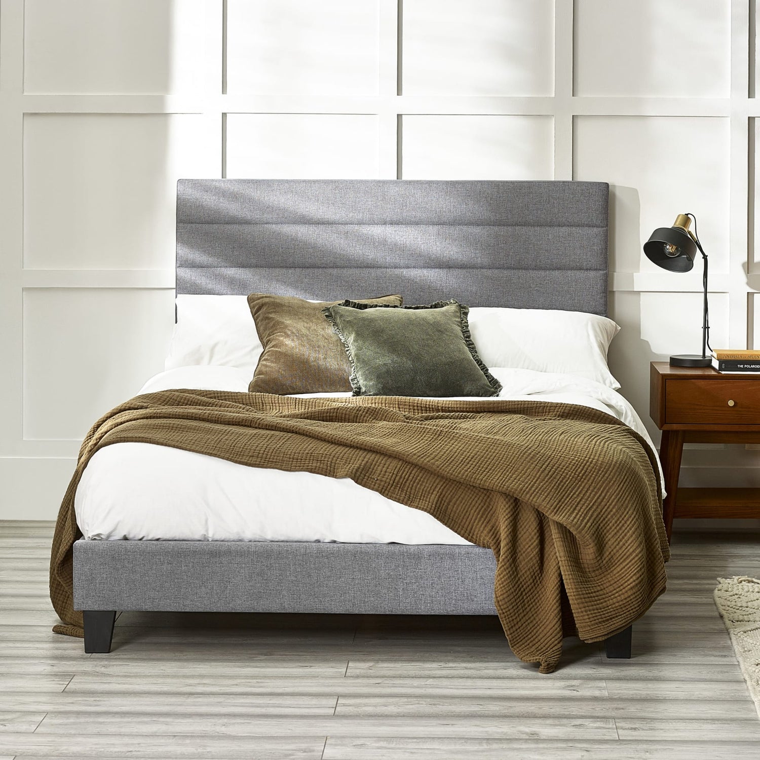 Gray upholstered bed with brown blanket and pillows against a white paneled wall.