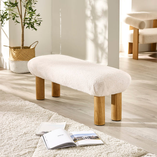 White cushioned bench with wooden legs in a room with a plant and open book on the floor.