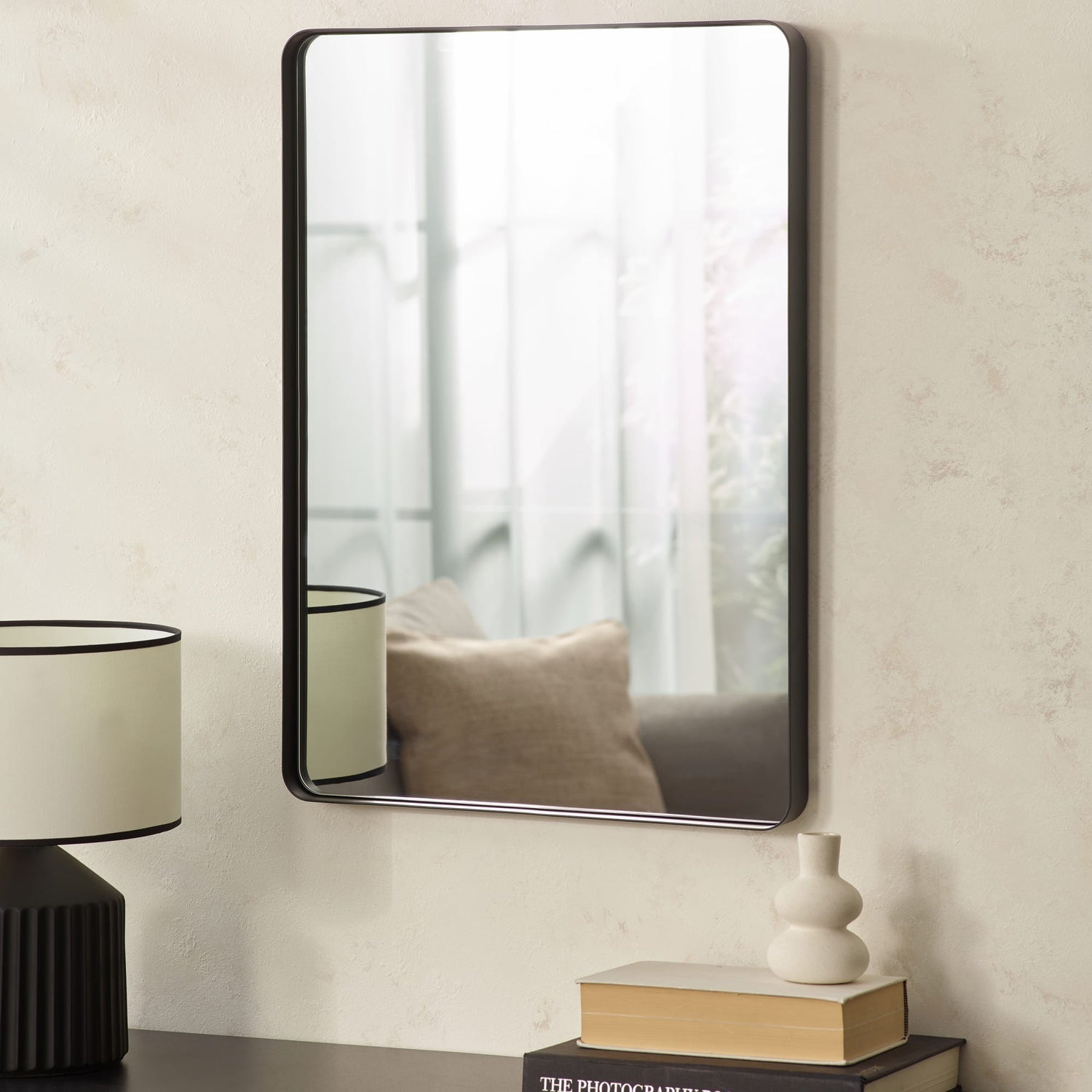 Rectangular mirror with black frame on a wall above books and a lamp.